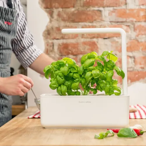 Click and Grow Smart Garden 3 in Weiß, rechteckiger Pflanzkübel mit L-förmigem LED-Arm und üppigem Basilikum auf einer hölzernen Küchenarbeitsplatte.