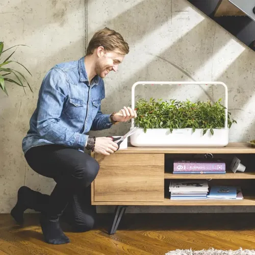 Smart Garden 9 Pro, vit rektangulär inomhusträdgård med bågformad ljusarm över tät grönska, på träbänk; person klipper.