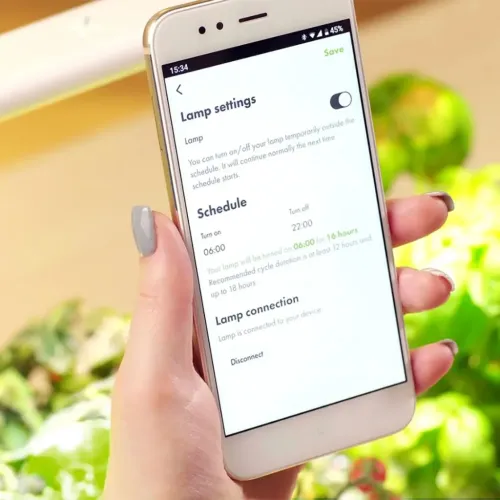 Smart Garden 9 Pro-appen visas på en smartphone med Lamp settings och schemaläggning, hållen i hand framför oskarpa växter.